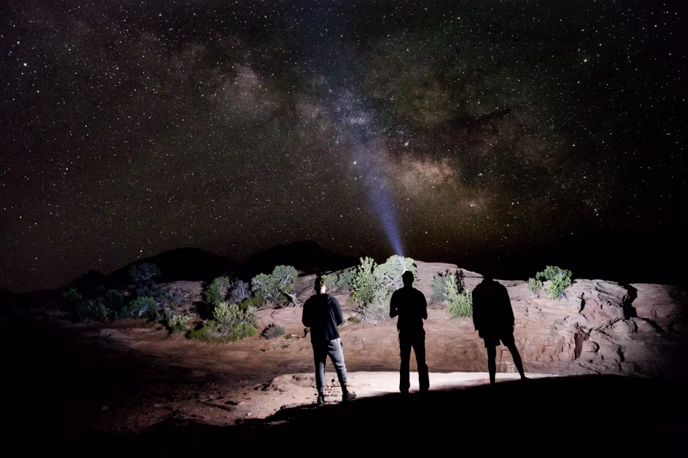 Nachtelijke buitenfoto met drie figuren op rots, lichtstraal naar hemel, heldere melkweg sterrenhemel, thema sterrenkijken natuur