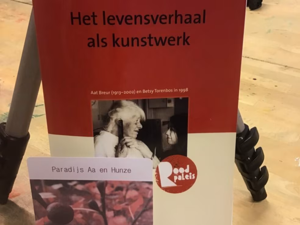 Poster met titel Het levensverhaal als kunstwerk, verwijzing naar Aat Breur 1913-2002 en Betsy Torenbos 1998, zwart-witfoto van gesprek, logo Pod pates, kaart Paradijs Aa en Hunze, houten vloer en metalen driepootstatief zichtbaar