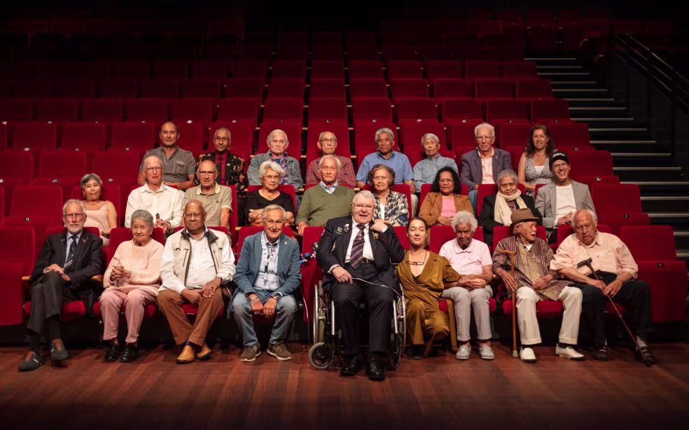 Groepsfoto in theater met 22 mensen zittend en staand over drie rijen rode stoelen. sfeer van bijeenkomst