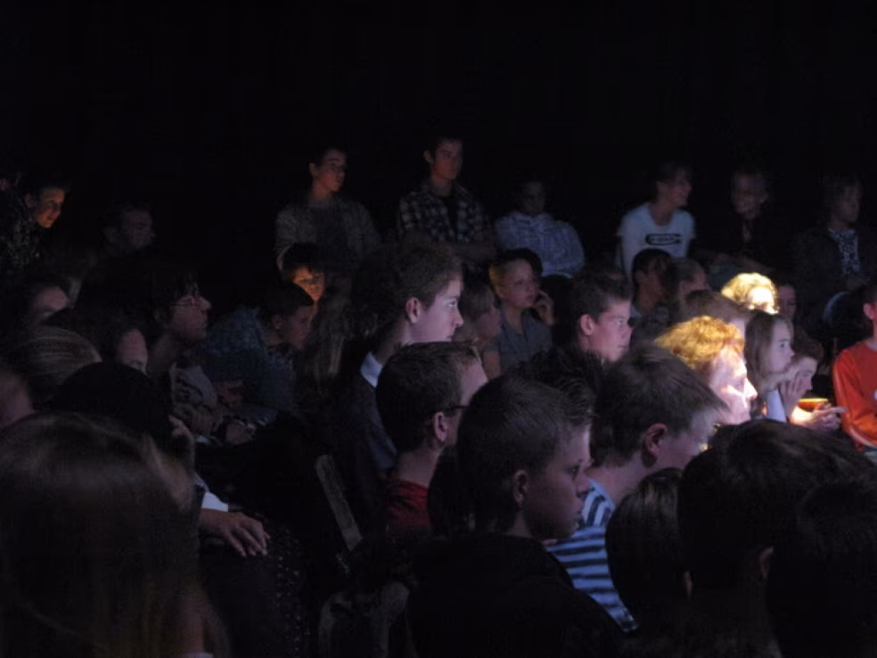 Groep mensen geconcentreerd kijken naar voorstelling in donkere ruimte met licht op gezichten